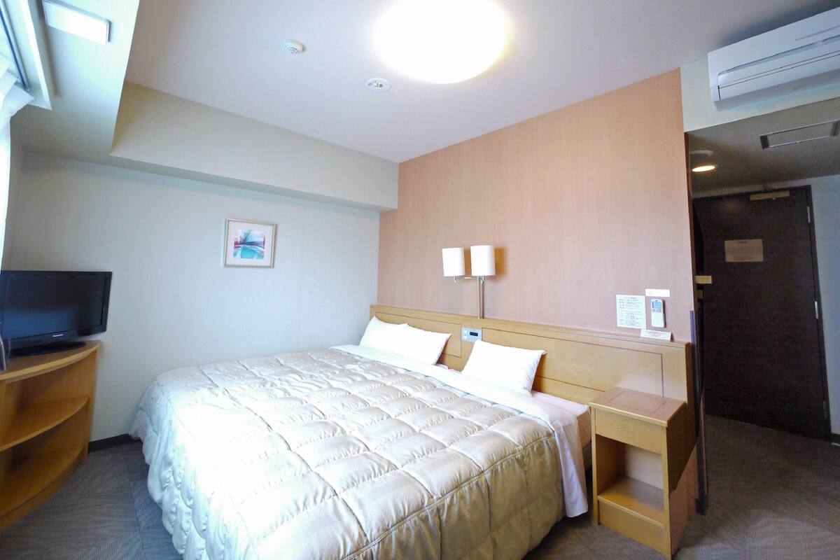 A hotel room with a bed, bedside tables, a television, wall-mounted lamps, and an air conditioning unit.