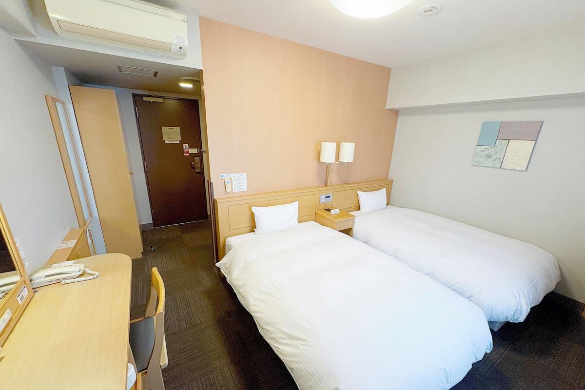 A hotel room with two single beds, a desk with a chair, a telephone, and a wall-mounted lamp.