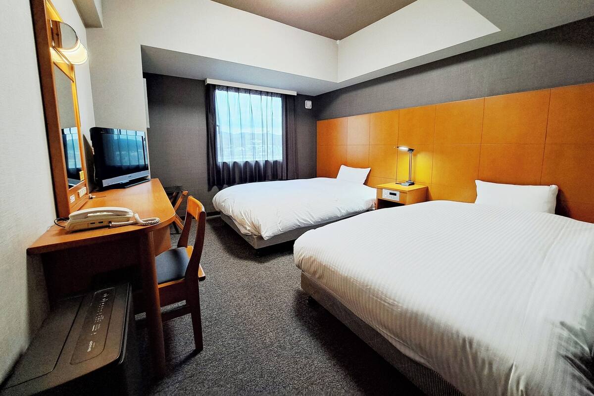 A hotel room with two beds, a desk with a telephone, a chair, a TV, and a window with curtains.