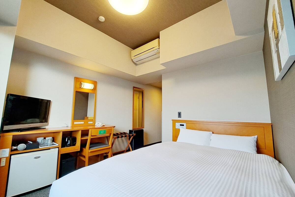 A hotel room with a bed, a desk with a television, a mirror, and a small refrigerator.