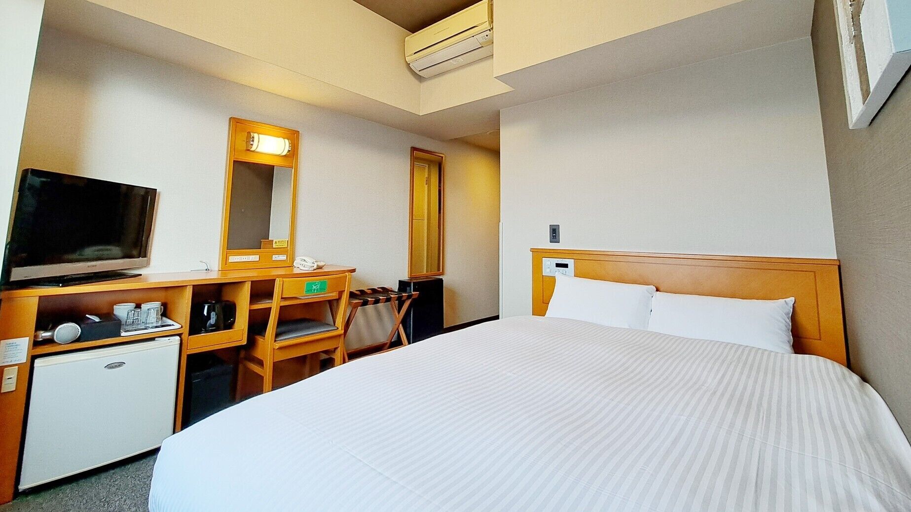 A hotel room with a bed, a desk with a TV, a small fridge, a mirror, and a door.