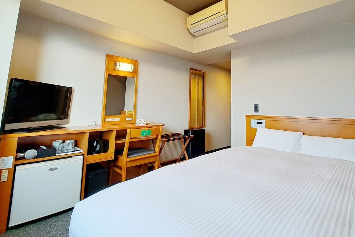 Une chambre d’hôtel avec un lit, un bureau avec une télévision, un petit réfrigérateur, un miroir et une porte.