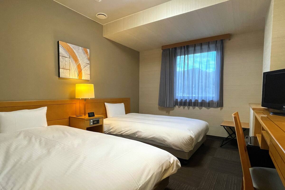 Une chambre d’hôtel avec deux lits, un bureau avec une télévision, une fenêtre avec des rideaux et un tableau au mur.