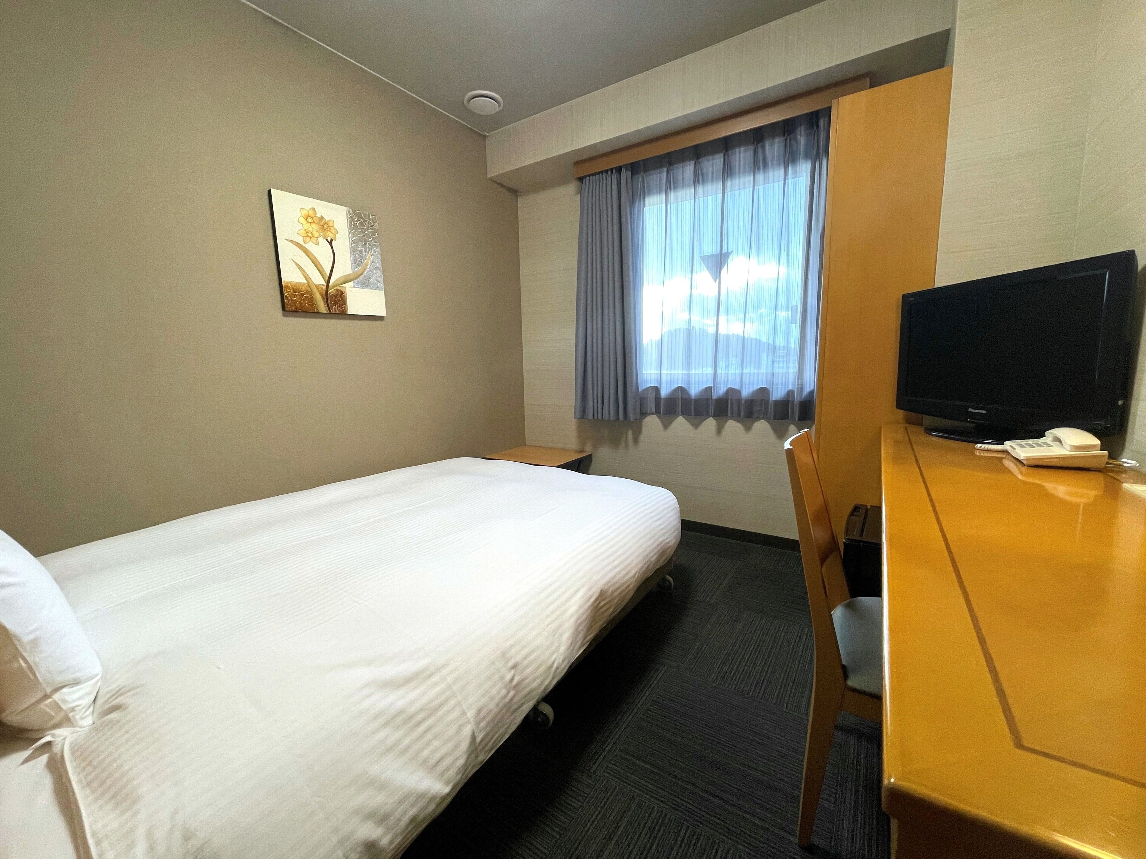 A hotel room with a single bed, a desk with a television, a telephone, and a window with curtains.