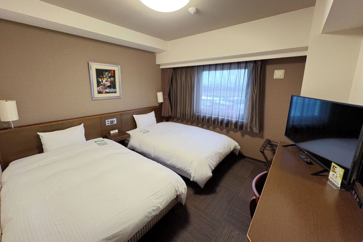 A hotel room with two beds, a television, a desk, and a window with curtains.