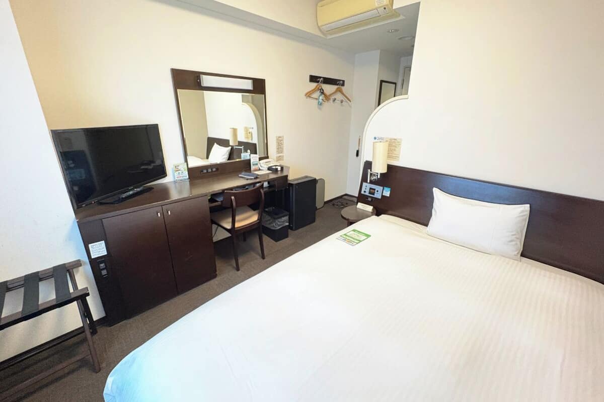 A hotel room with a large bed, a desk with a chair, a television, a mirror, and a bedside table with a lamp.