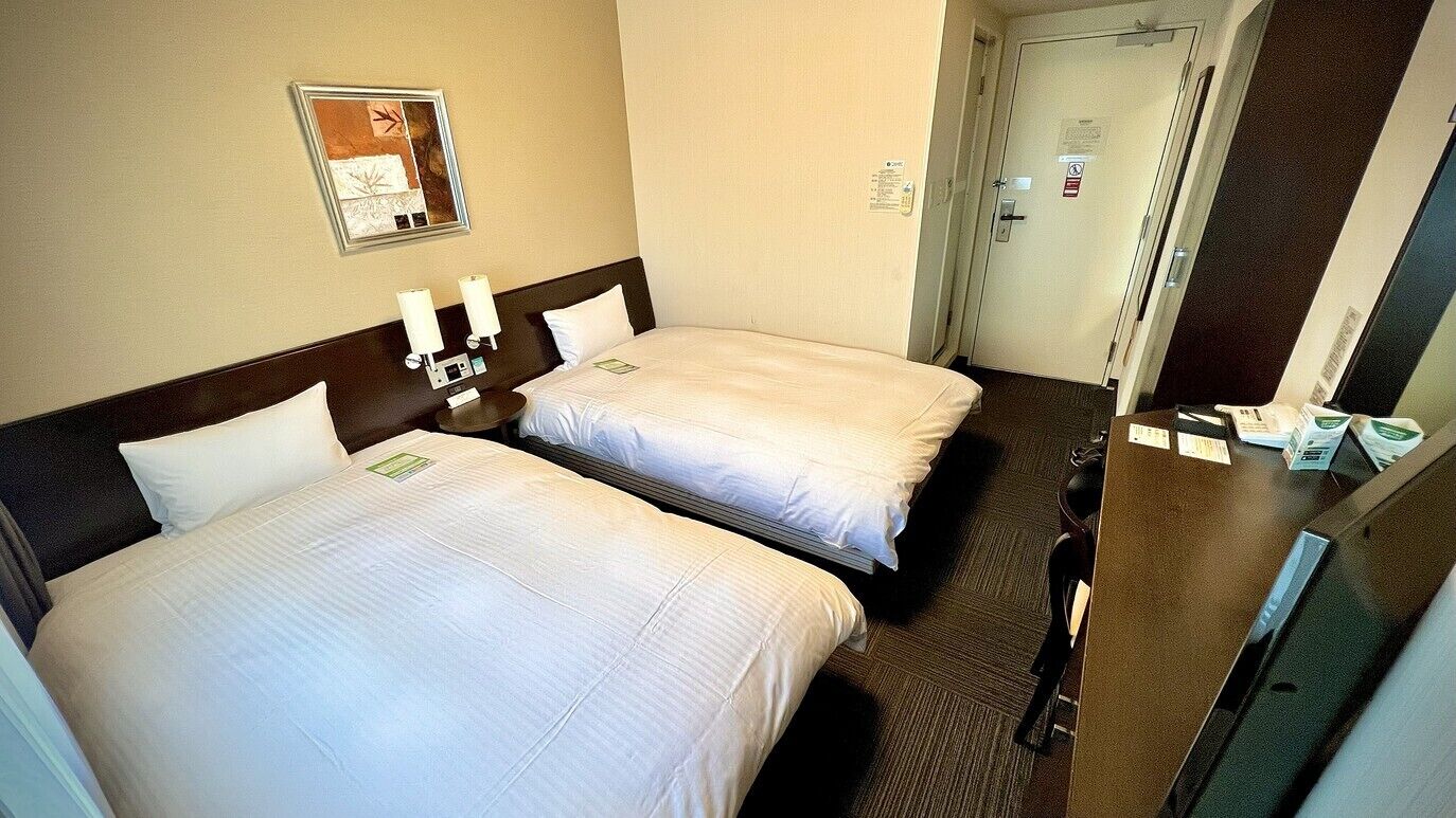 Una habitació d'hotel amb dos llits, una tauleta de nit, una làmpada penjada a la paret i una porta amb un cartell.