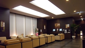 Un área de recepción con una hilera de sofás color beige, un mostrador de recepción con computadoras y un ventanal con persianas.