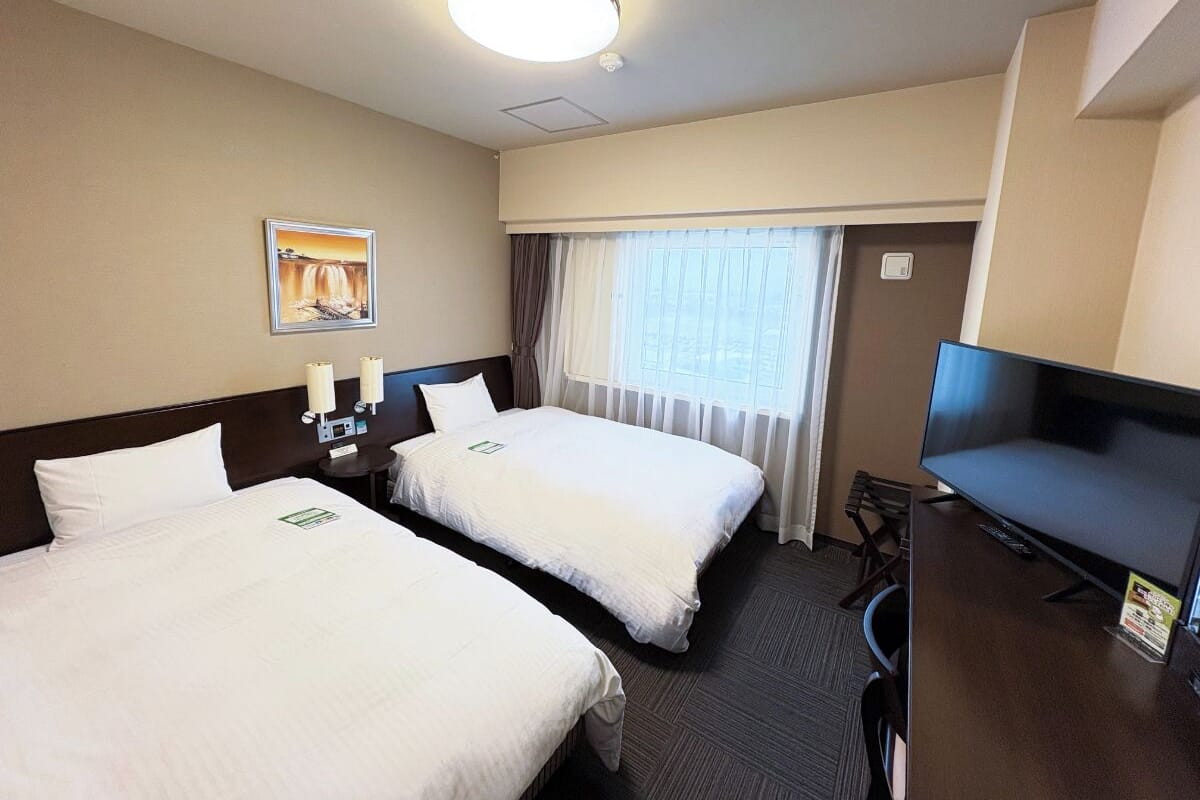 A hotel room with two beds, a desk with a television, a window with curtains, and a painting on the wall.