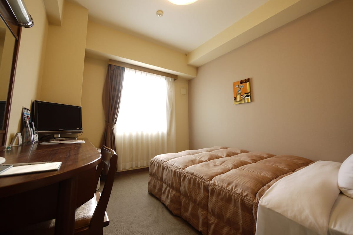 A hotel room with a bed, a desk with a computer, a chair, and a window with curtains.