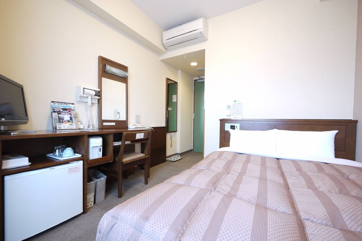 A hotel room with a bed, a desk, a chair, a television, a mini-fridge, and an air conditioning unit.