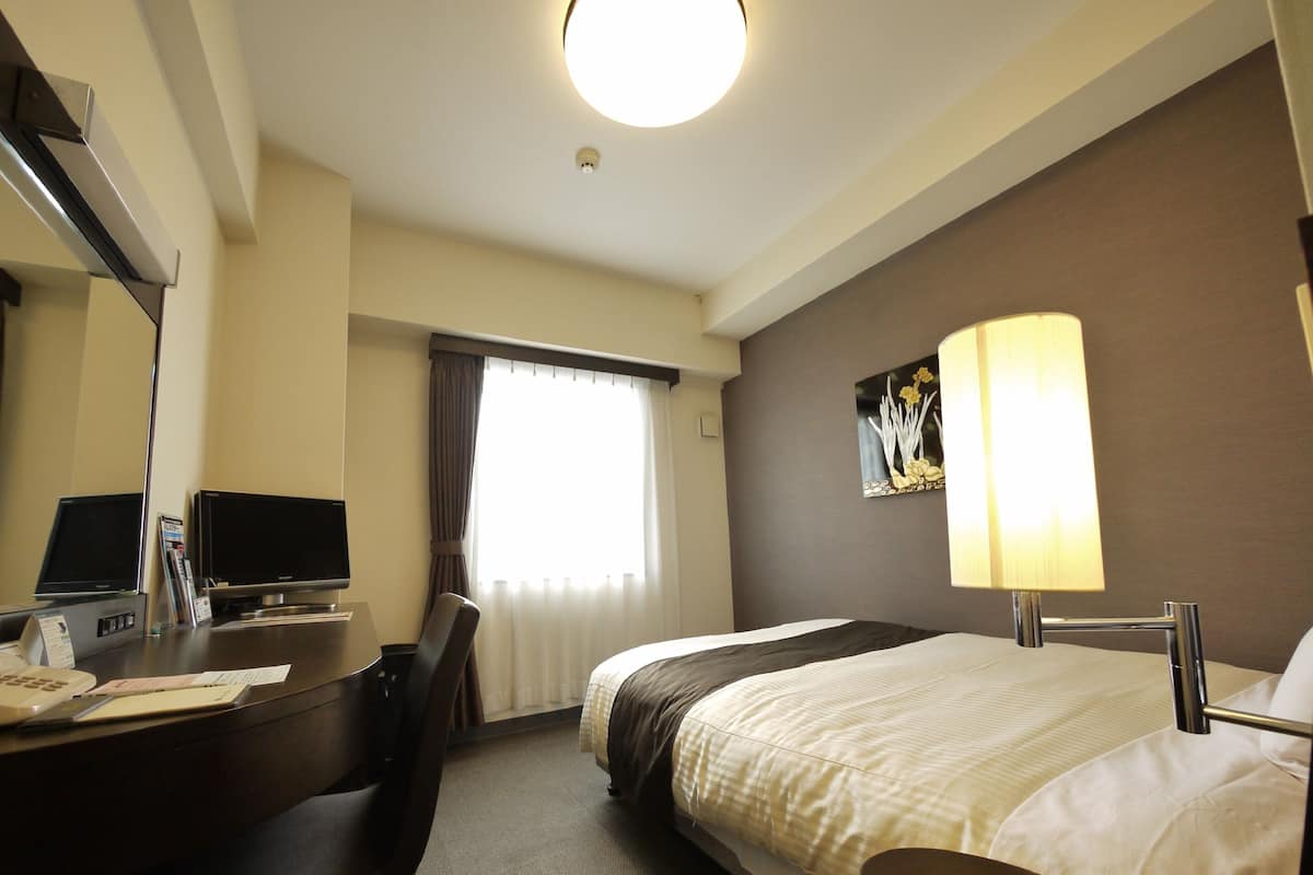A hotel room with a bed, desk, chair, television, and a lamp.