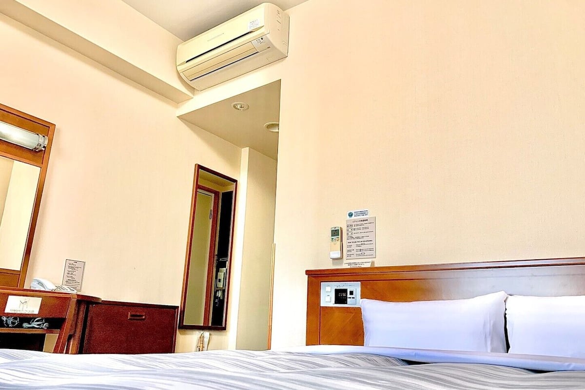Quarto de hotel com cama, escrivaninha, espelho, aparelho de ar condicionado e painel de controle fixado na parede.