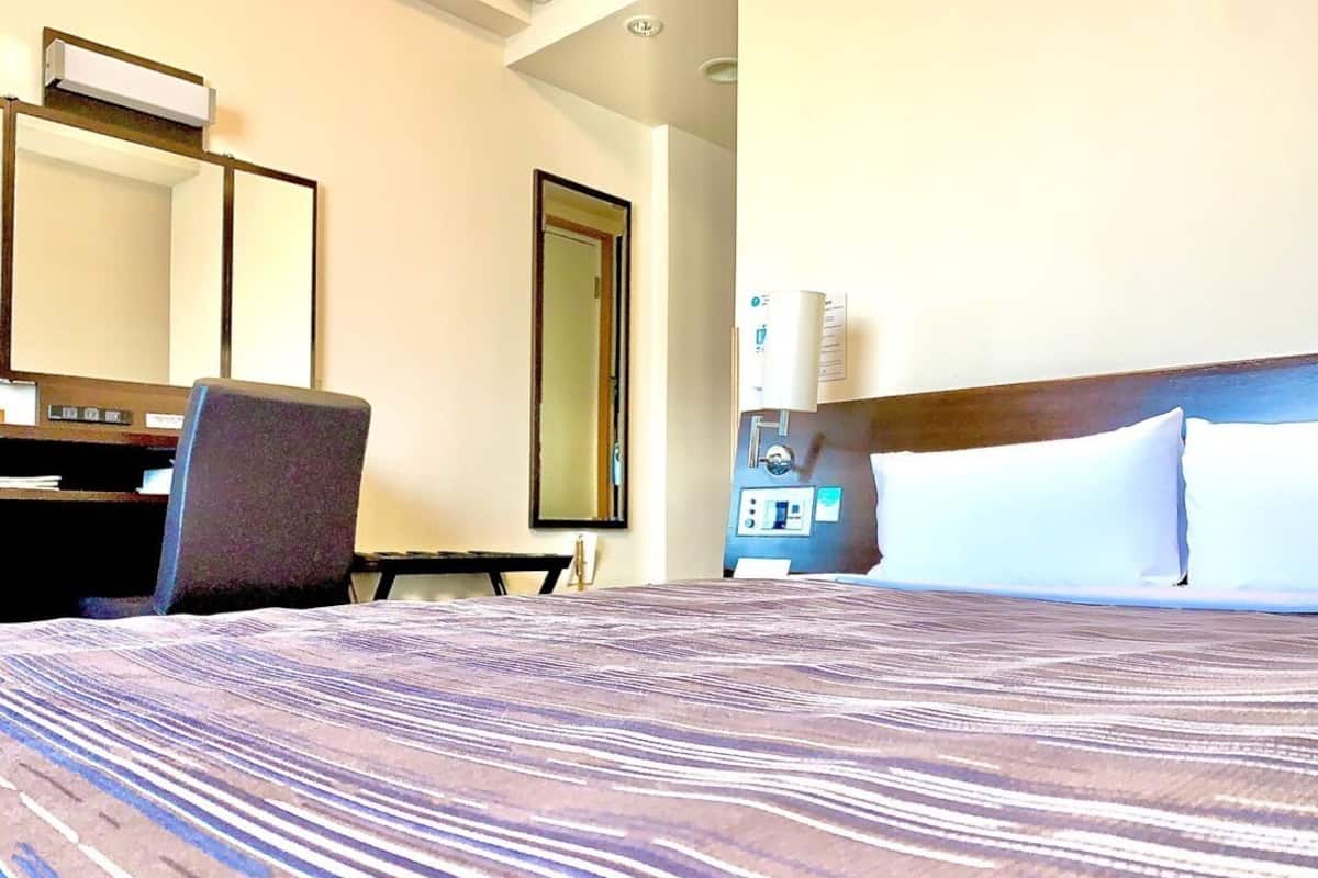 Quarto de hotel com cama, escrivaninha, cadeira, espelho e mesa de cabeceira com abajur.