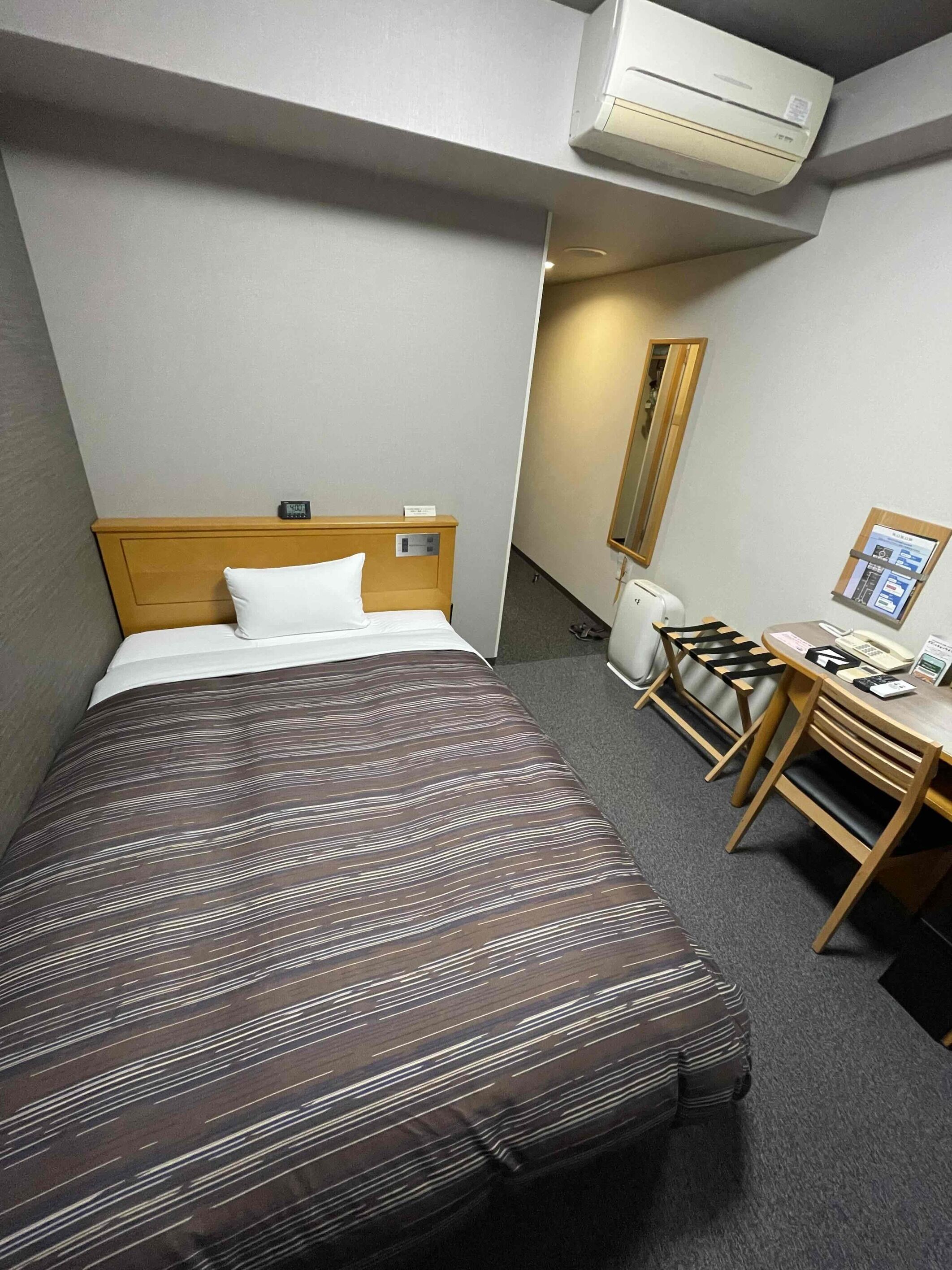 A hotel room with a bed, a desk, a chair, an air conditioner, and a mirror.