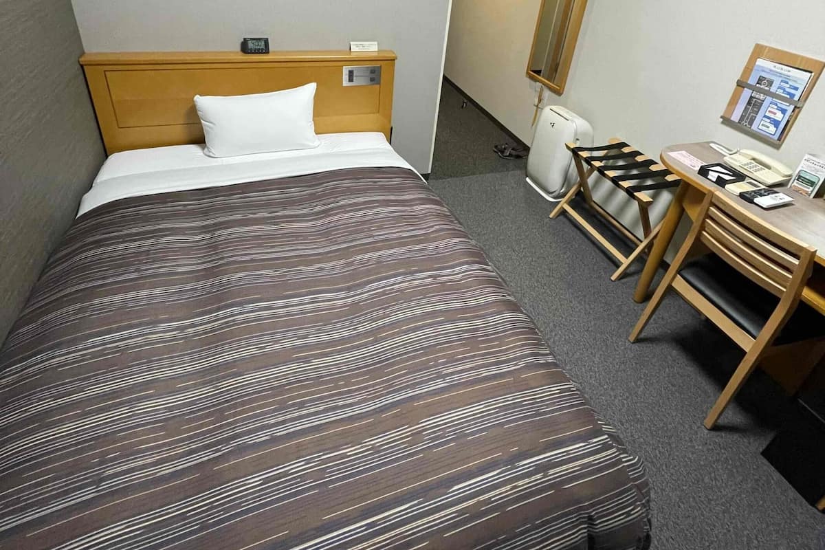 A hotel room with a bed, a desk, a chair, an air conditioner, and a mirror.