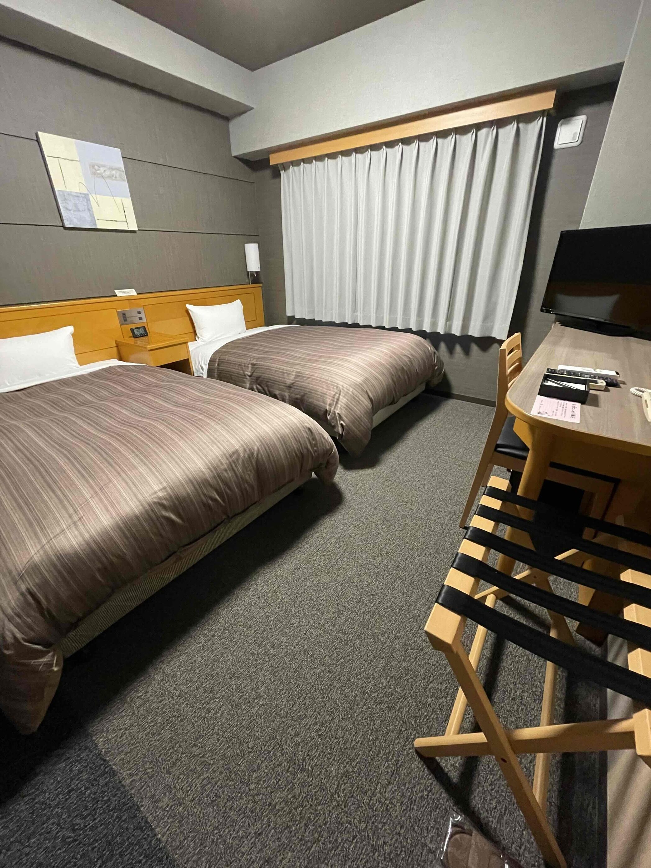 A hotel room with two beds, a desk with a chair, a television, and a window with curtains.