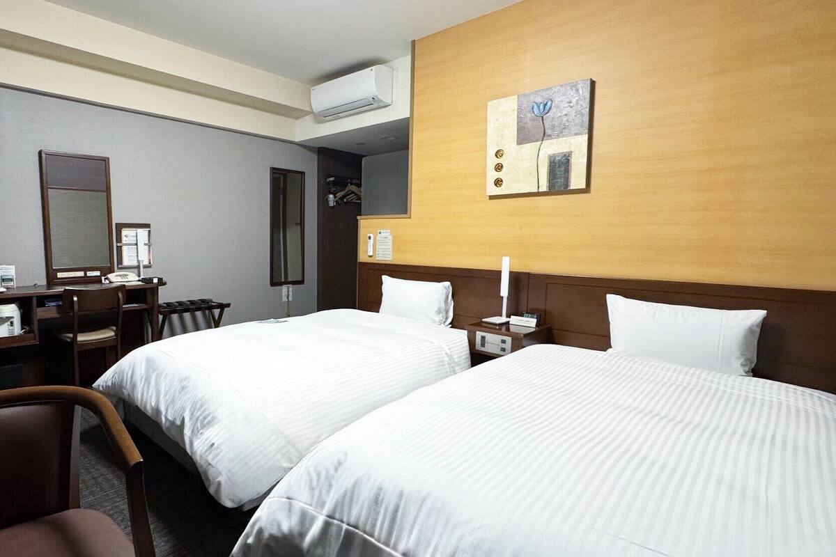 A hotel room with two beds, a desk, a chair, an air conditioning unit, and a painting on the wall.