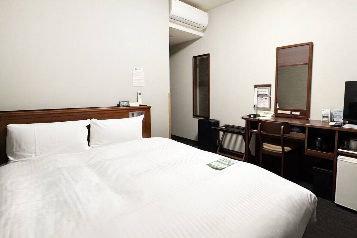 A hotel room with a bed, a desk, a chair, a mirror, a television, and an air conditioning unit.