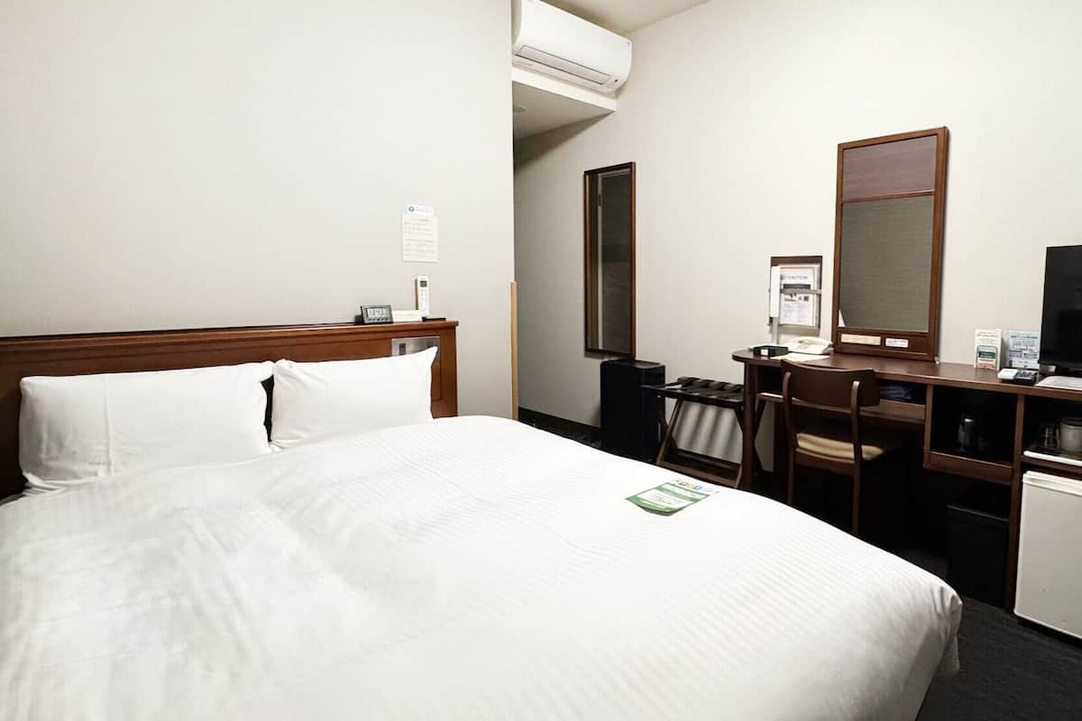 A hotel room with a bed, a desk, a chair, a mirror, a television, and an air conditioning unit.