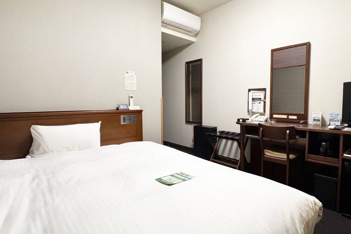 A hotel room with a bed, a desk, a chair, a mirror, an air conditioning unit, and a bulletin board.