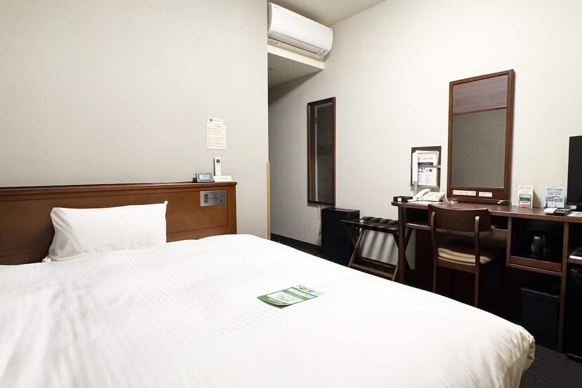A hotel room with a bed, a desk, a chair, a mirror, an air conditioning unit, and a bulletin board.