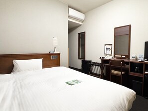 Ett hotellrum med en säng, ett skrivbord, en stol, en spegel, en luftkonditioneringsanläggning och en anslagstavla.