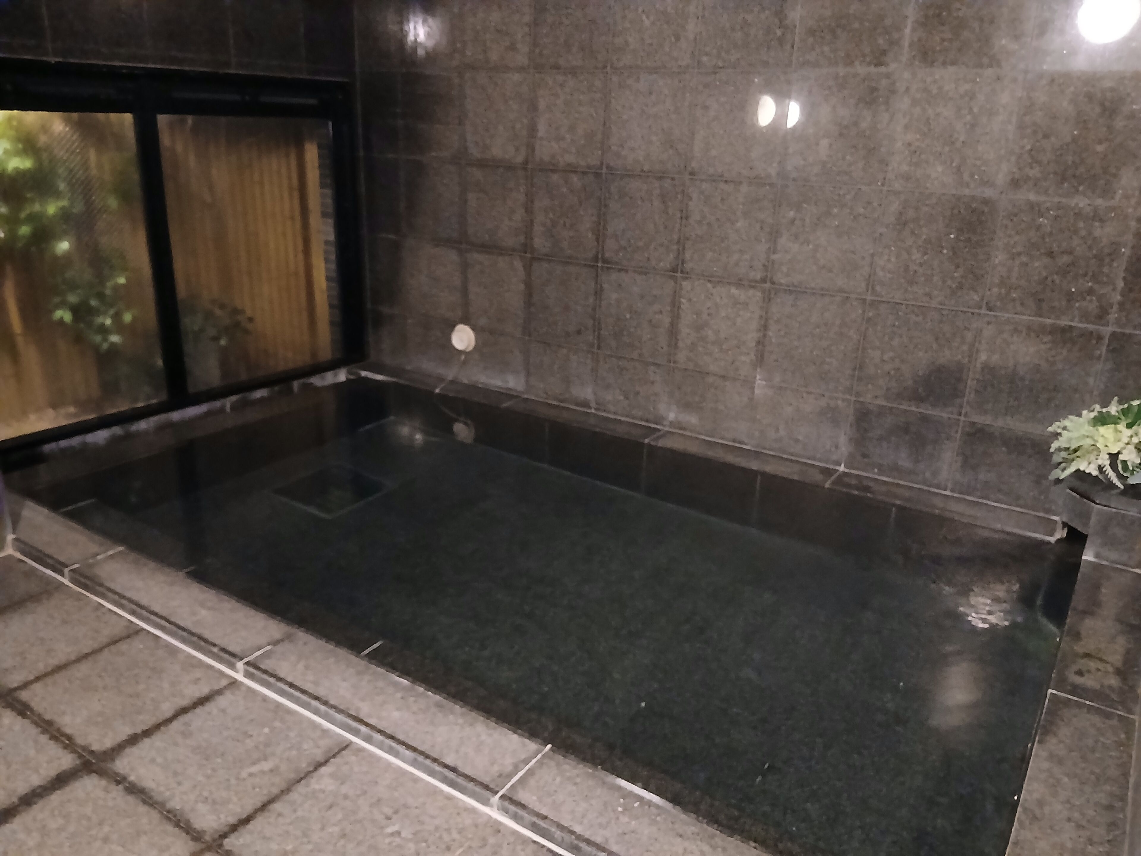 Een rechthoekig bubbelbad met een donker, spiegelend oppervlak, omringd door donkere tegels.