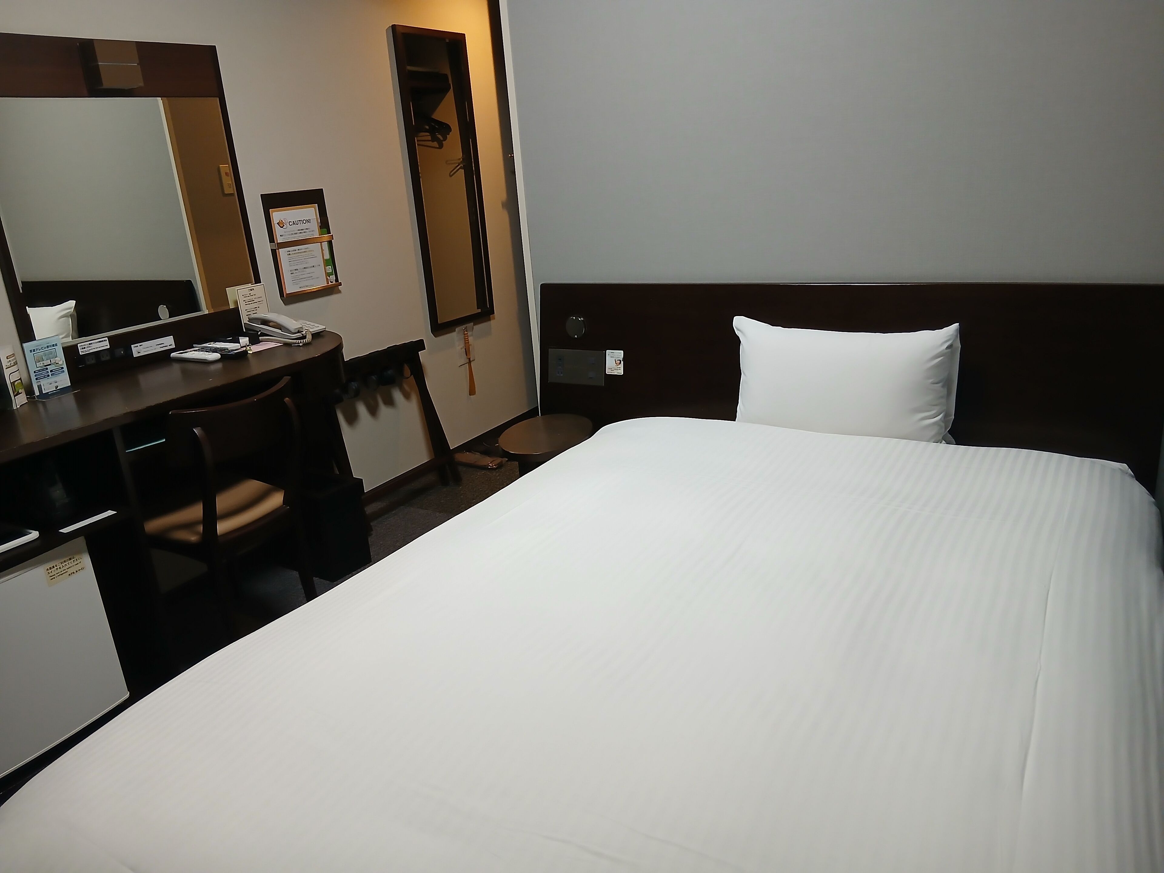 Una habitación de hotel con cama, escritorio, silla, espejo y perchero.