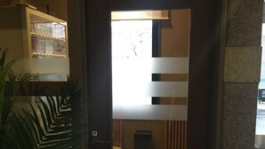 A hotel room entrance with a wooden door, a potted plant, and a glimpse of a seating area with a trash bin and a cabinet. - Hotel Route Inn Tosu Ekimae (Tosu)