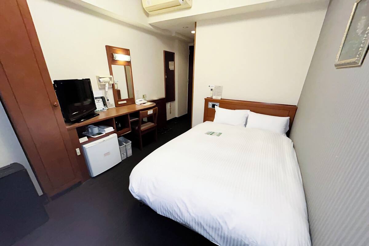 A hotel room with a bed, a desk with a computer, a chair, a small refrigerator, and a television.