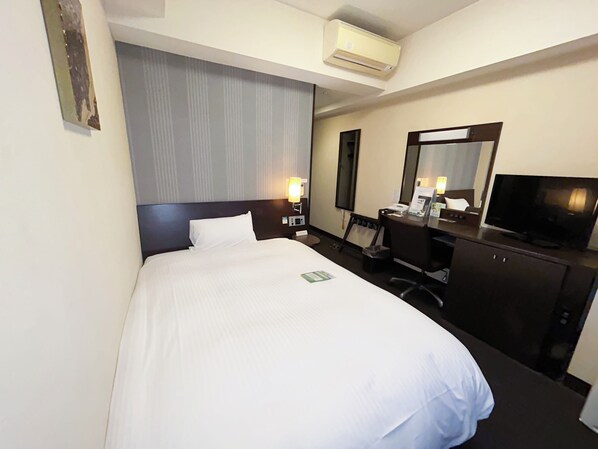 A hotel room with a bed, a desk with a computer, a television, and an air conditioning unit.