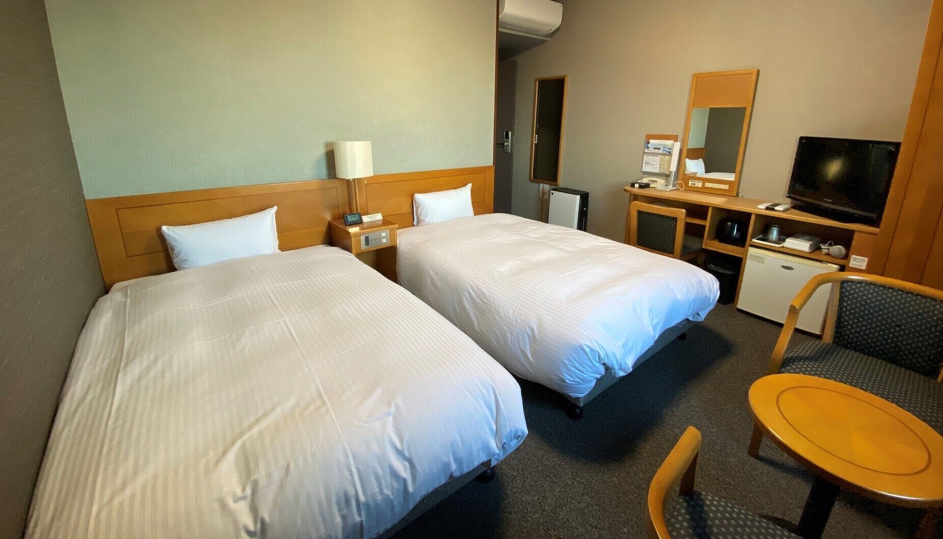 A hotel room with two beds, a desk, a chair, a television, and a small kitchenette.