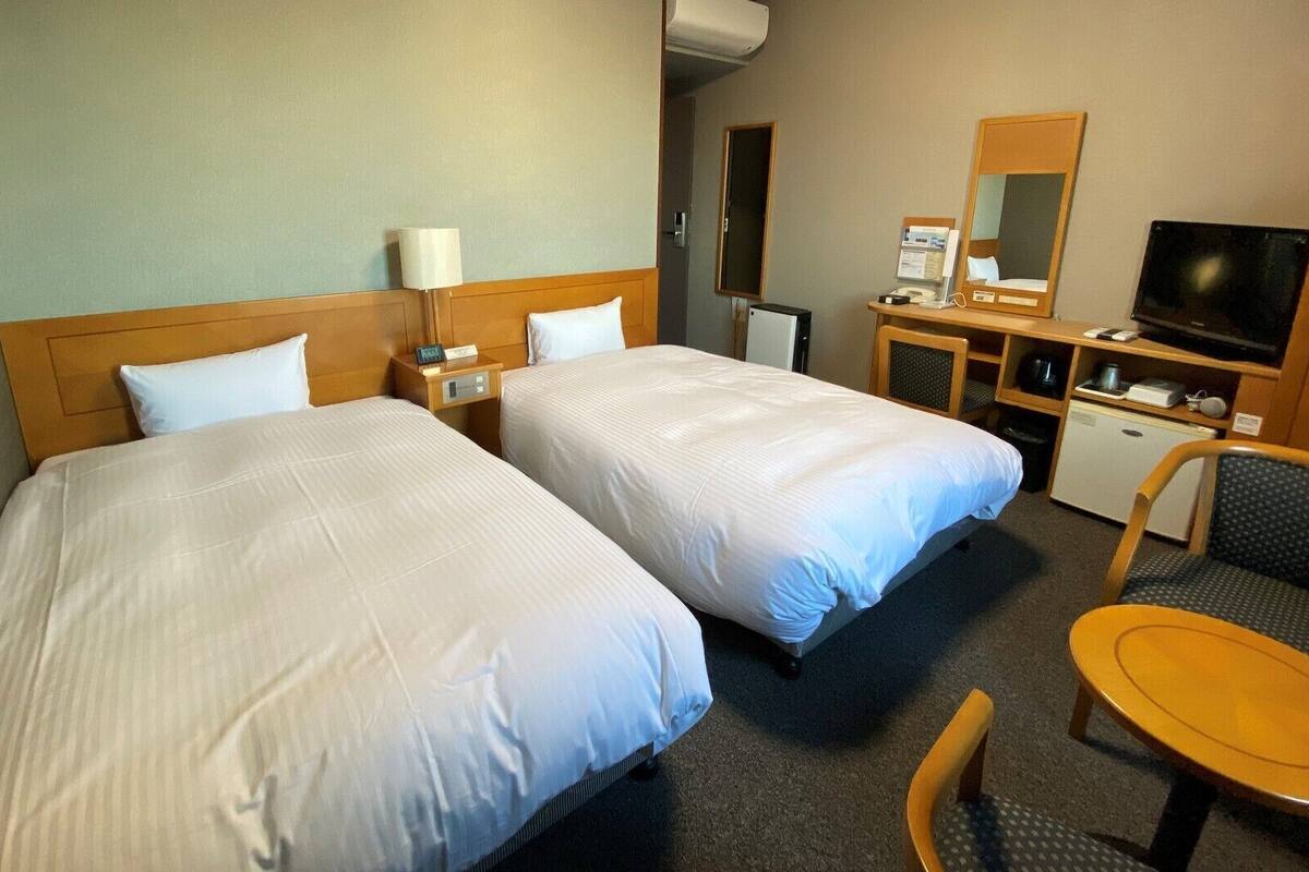 A hotel room with two beds, a desk, a chair, a television, and a small kitchenette.