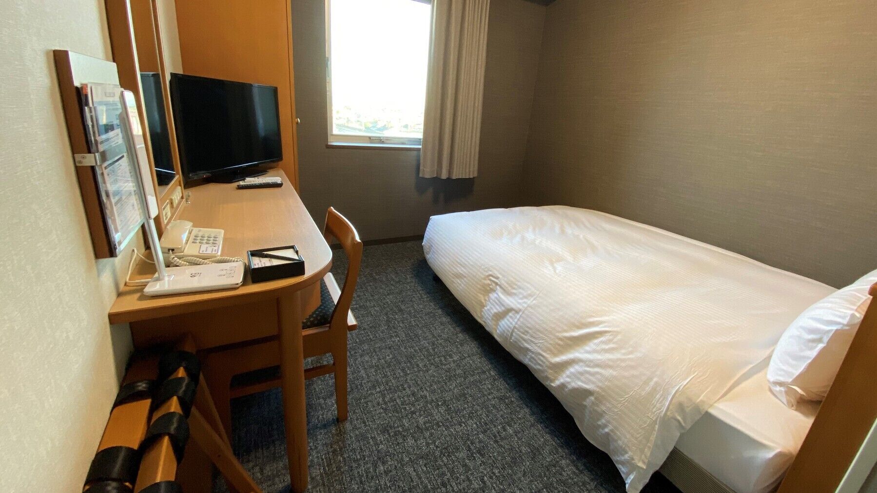 A hotel room with a single bed, a desk with a computer, a telephone, and a chair.