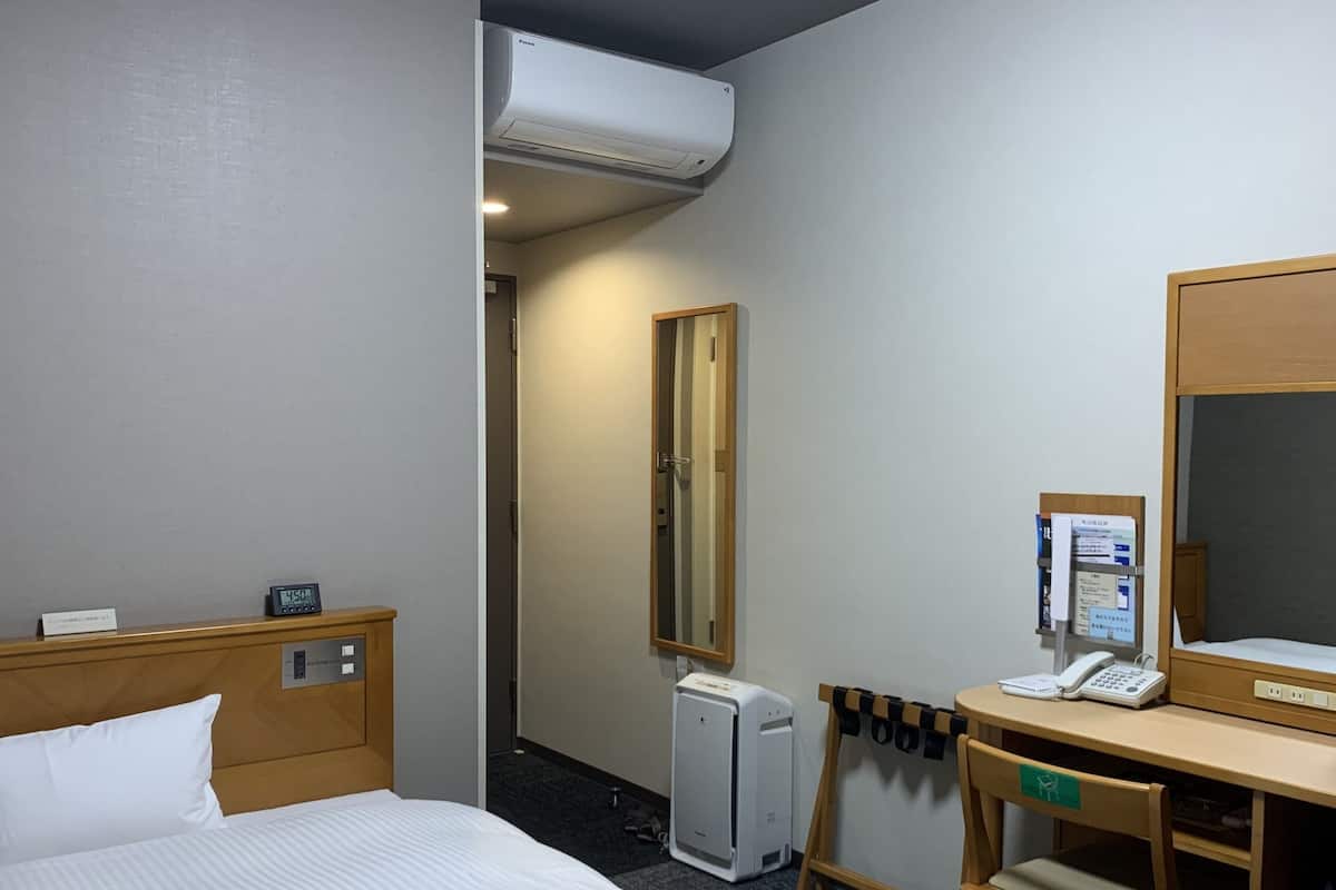 A hotel room with a bed, a desk with a telephone, a mirror, and a small heater.