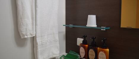 A bathroom with a white towel hanging on a rack, a glass shelf with a white cup, and three bottles of body soap and shampoo.