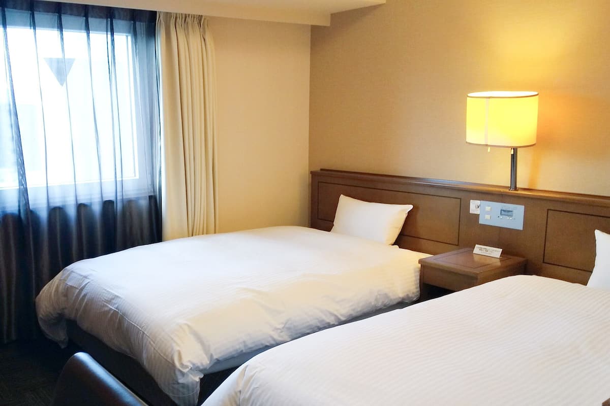 A hotel room with two beds, a nightstand, a lamp, and a window with curtains.