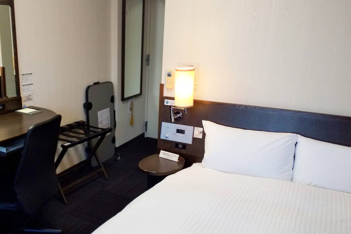 Une chambre d’hôtel équipée d’un lit, d’un bureau, d’une chaise, d’une table de chevet et d’une lampe fixée au mur.