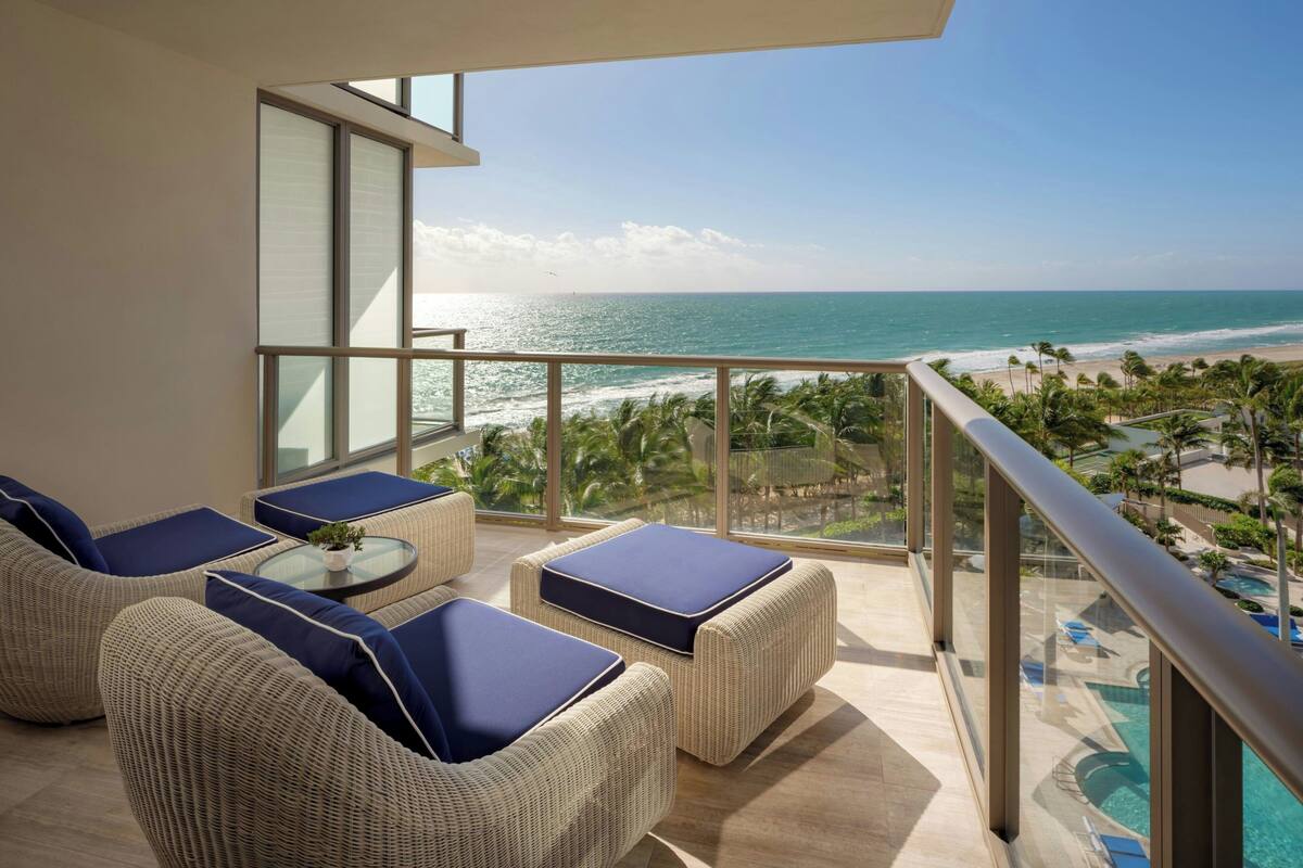 Un balcón con mobiliario de mimbre y barandilla de vidrio con vistas a una playa y una zona de piscina.