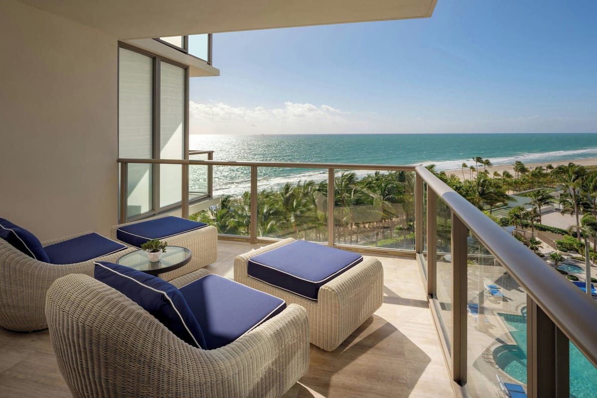 A balcony with wicker furniture and a glass railing overlooking a beach and pool area.