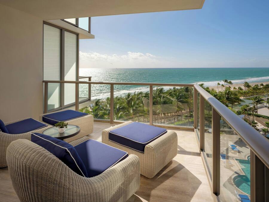 A balcony with wicker furniture and a glass railing overlooking a beach and pool area.