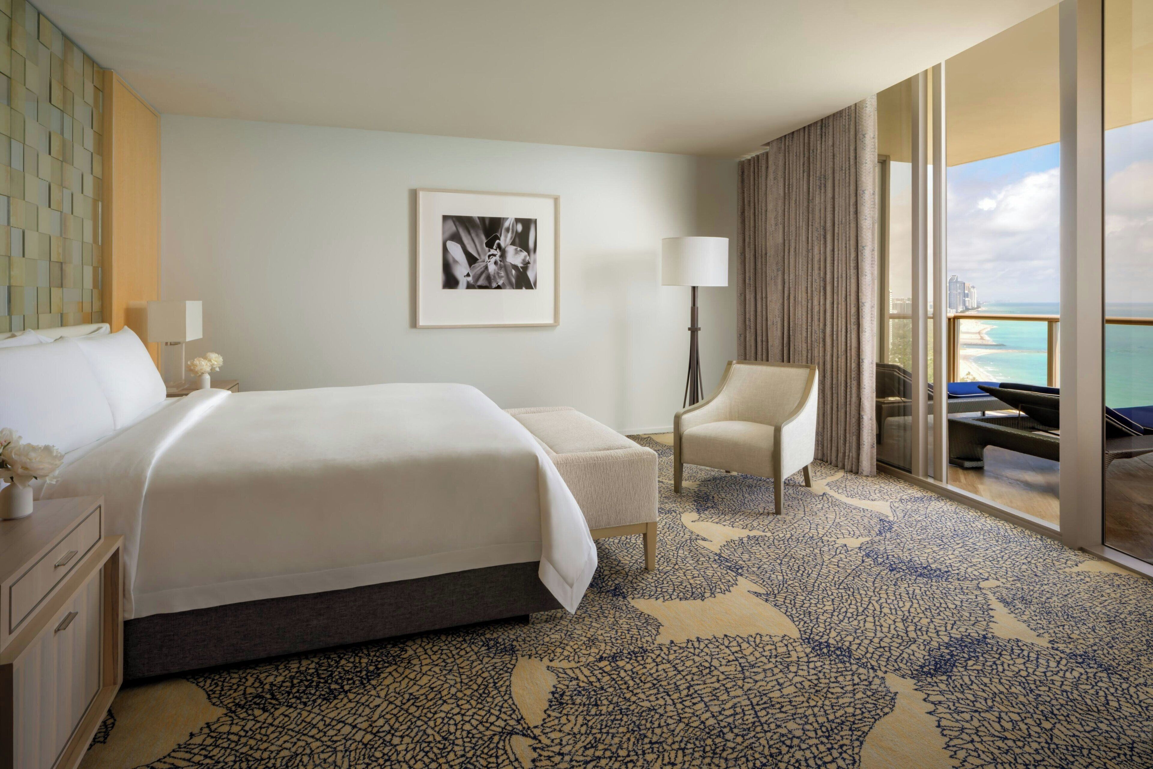 A hotel room with a large bed, a chair, a desk, a lamp, a framed picture, and a balcony with a view of the ocean.