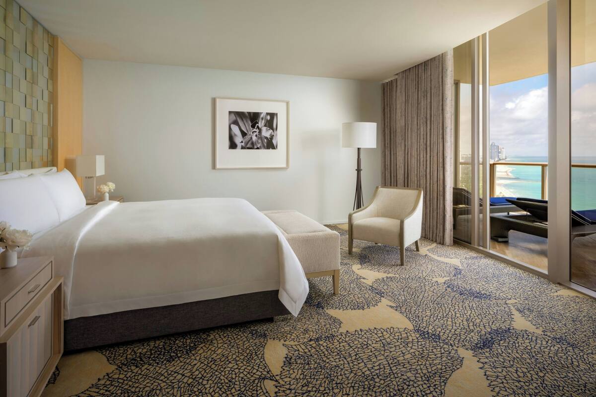 Habitación de hotel con una cama grande, una silla, un escritorio, una lámpara, un cuadro enmarco y un balcón con vista al océano.