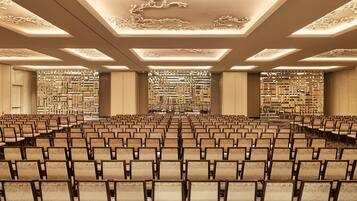 Ein großer Konferenzraum mit Stuhlreihen, aufwändigen Deckenverzierungen und einer zentralen Wand mit Dekorationen.