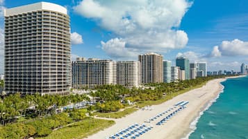 A beachfront hotel with a curved building, a sandy beach with rows of blue umbrellas, and a clear blue sky.