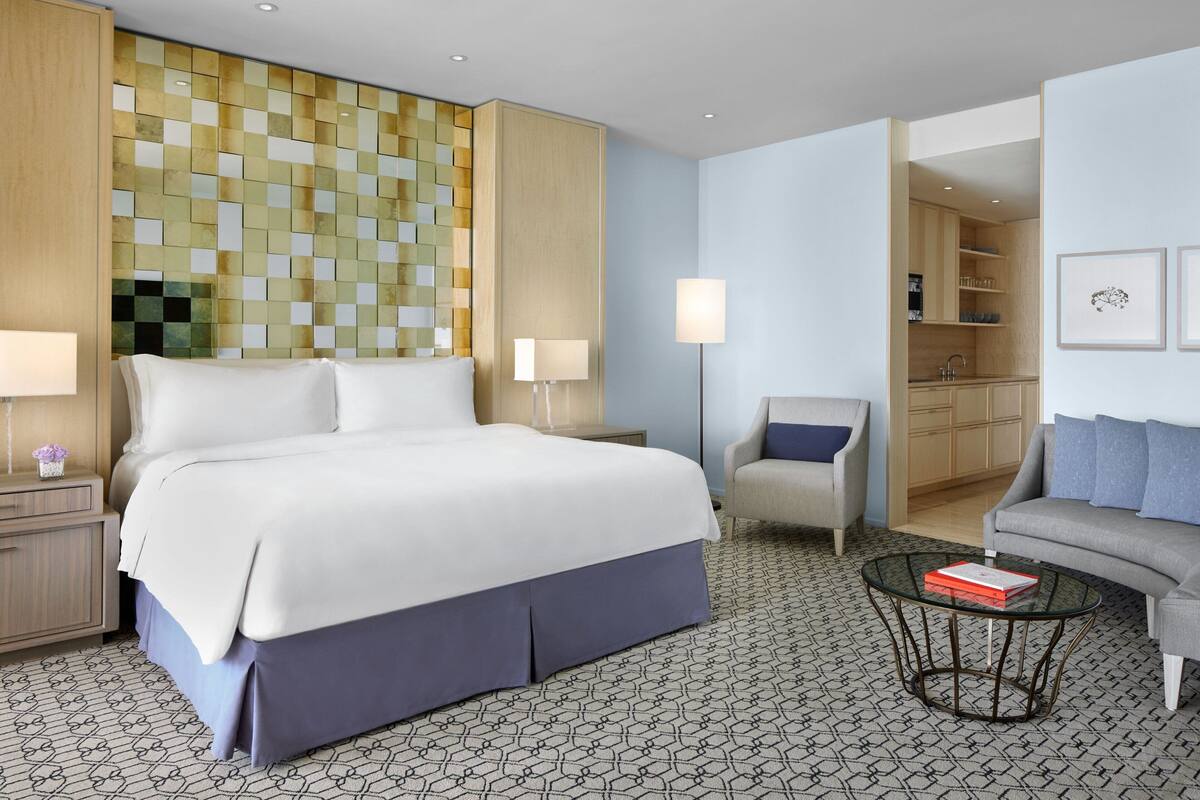Habitación de hotel con una cama grande, una zona de estar con sofá y sillón, una mesita y una kitchenette al fondo.