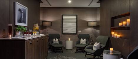 A modern spa room with a wooden reception desk, two armchairs, a small round table with a candle, and wall-mounted lamps.