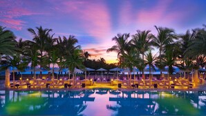 Un espace aménagé au bord de la piscine, avec des chaises longues et des parasols, entouré de palmiers, au coucher du soleil.