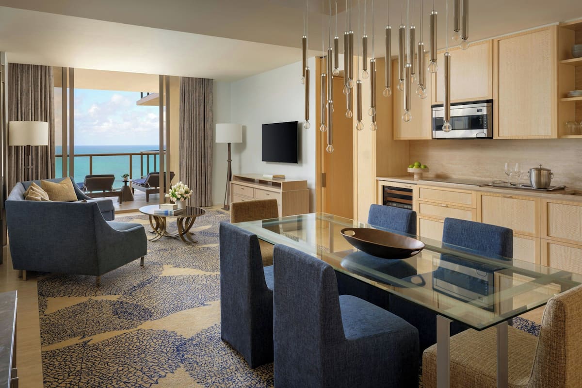 Una habitación de hotel moderna con balcón, zona de comedor con mesa de vidrio y cocina con armarios de madera.